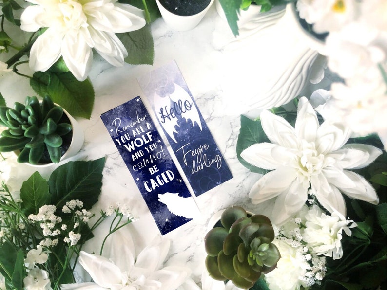 May include: Two blue and white bookmarks with a galaxy background. One bookmark features a wolf silhouette and the text "Remember you are a wolf and you cannot be caged". The other bookmark features a bat silhouette and the text "Hello Feyre darling".