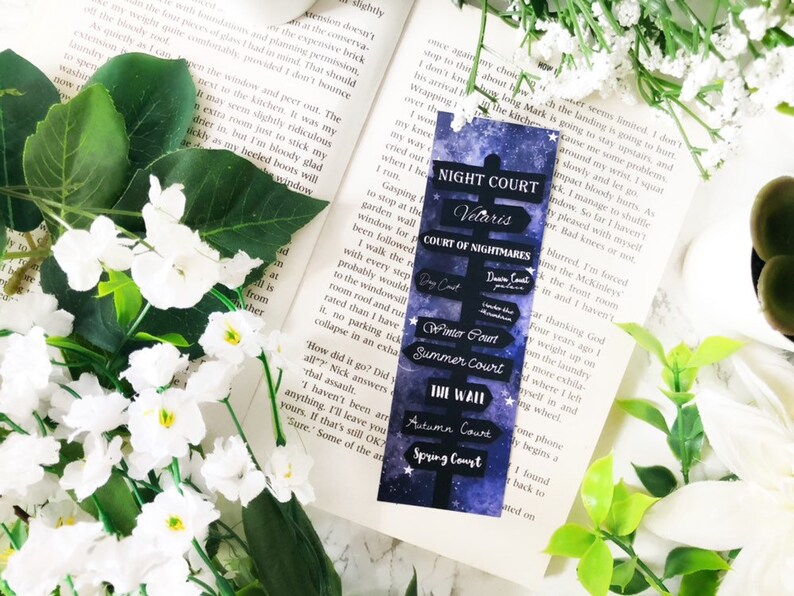 May include: A blue and purple bookmark with a starry background and a black sign that reads "Night Court Velaris Court of Nightmares" with arrows pointing to other courts: "Spring Court", "Autumn Court", "The Wall", "Summer Court", "Winter Court", "Dawn Court", and "Day Court".