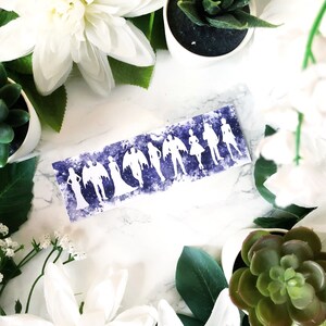May include: A white card with a purple watercolour background and white silhouettes of people. The card is surrounded by white flowers and green succulents.