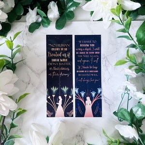 May include: Two bookmarks featuring quotes from a book. The bookmarks are dark blue with gold lettering and a silhouette of a woman in a dress. The first bookmark reads "No human ought to be treated as if their work didn't matter, or their choices, or their dreams.". The second bookmark reads "If I have to follow you across a desert, I will have to jump into the Nile again, and again. I will. If I have to leap in front of a thousand bullets I will. I will always love you."