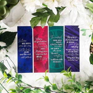 Może przedstawiać: Cztery kolorowe zakładki z cytatami z książki "Szklana Księżniczka" autorstwa Sarah J. Maas. Zakładki mają niebieski, czerwony, zielony i fioletowy kolor z białym tekstem.