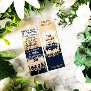 Peut inclure: Deux marque-pages rectangulaires avec des motifs dorés et bleu marine. L'un des marque-pages indique "THIS IS HOW IT STARTED" avec du texte supplémentaire. L'autre marque-page indique "LIBERTY EQUALITY" et "THE COURT OF MIRACLES". Les deux ont des bordures décoratives.