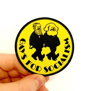 May include: A yellow circular patch with black text that reads "GAYS FOR SOCIALISM". Two figures in black are walking side-by-side on the patch.