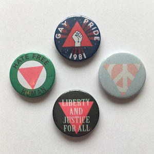 May include: Four round buttons with different designs and messages. The first button is green with a pink triangle and the text "Hate Free Human". The second button is blue with a red fist in a triangle and the text "Gay Pride 1981". The third button is light blue with a pink peace sign in a triangle. The fourth button is black with a pink triangle and the text "Liberty and Justice for All".