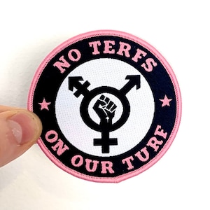 Może przedstawiać: Naszywka haftowana w kolorze różowym i czarnym z tekstem "NO TERFS ON OUR TURF" i symbolem transgender z uniesioną pięścią w środku.