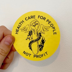 May include: Yellow round sticker with black text that reads "Health Care For People Not Profit". A black and white illustration of a fist with wings and a dragon wrapped around it is in the center.