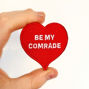 May include: Red felt heart-shaped patch with white embroidered text that reads "BE MY COMRADE".