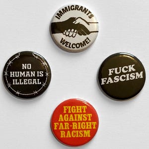 Könnte beinhalten: Vier runde Buttons mit politischen Botschaften. Ein Button zeigt "IMMIGRANTS WELCOME!" mit einer Handshake-Grafik. Ein anderer Button zeigt "NO HUMAN IS ILLEGAL" mit Stacheldraht. Ein dritter Button zeigt "FUCK FASCISM". Der letzte Button zeigt "FIGHT AGAINST FAR-RIGHT RACISM".
