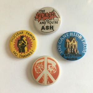 May include: Four round buttons with different designs. The first button is yellow with a red sun and a silhouette of a family. The text reads "Nuclear Family No Thanks!" The second button is white with a red and yellow graphic of a mushroom cloud and the text "One Flash And You're Ash". The third button is blue with a white house over a man's head and the text "You Can't Hide From A Neutron Bomb". The fourth button is orange with a white peace sign.