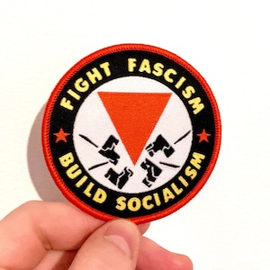 May include: A red, white, and black circular patch with the text "Fight Fascism Build Socialism" and a red triangle with a white star on each side. The triangle is broken by two white fists.