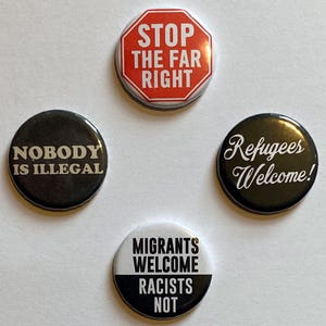 May include: Four round badges with political slogans. One black badge has white text reading "NOBODY IS ILLEGAL". Another says "Refugees Welcome!" in white script on black. A third badge says "MIGRANTS WELCOME RACISTS NOT" in black and white. The fourth says "STOP THE FAR RIGHT" in red and white.