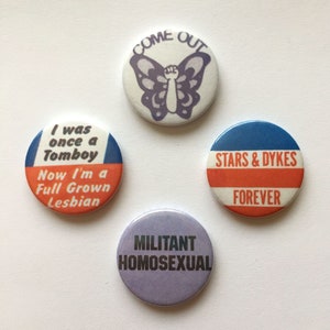 Può includere: Quattro bottoni rotondi con testo e grafica. Un bottone ha un disegno a farfalla e le parole "COME OUT". Un altro dice "I was once a Tomboy Now I'm a Full Grown Lesbian". Un terzo dice "STARS & DYKES FOREVER". L'ultimo dice "MILITANT HOMOSEXUAL".