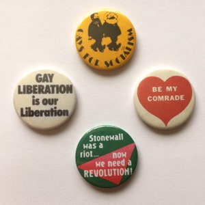Pode incluir: Quatro botões redondos com mensagens e designs diferentes. O botão superior é amarelo com texto preto que diz "Gays for Socialism". O botão esquerdo é branco com texto preto que diz "Gay Liberation is our Liberation". O botão direito é branco com um coração vermelho e texto preto que diz "Be My Comrade". O botão inferior é verde com texto vermelho e branco que diz "Stonewall was a riot... now we need a Revolution!"