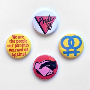 May include: Four round button pins with different designs. The first pin is white with a pink triangle and the text "Pride '85". The second pin is yellow with a blue double female symbol. The third pin is red with a pink and black handshake. The fourth pin is yellow with the text "We are the people our parents warned us against".