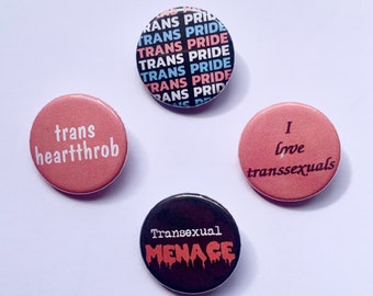 May include: Four round button badges with different messages. One badge is black with white text that reads "Trans Pride" repeated multiple times. Another badge is pink with white text that reads "trans heartthrob". A third badge is pink with black text that reads "I love transsexuals". The final badge is black with white text that reads "Transexual MENAGE" in dripping letters.