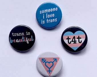 Könnte beinhalten: Vier runde Knöpfe mit verschiedenen Designs. Der erste Knopf ist blau mit weißem Text, der "someone i love is trans" lautet. Der zweite Knopf ist schwarz mit weißem Text, der "trans is beautiful" lautet. Der dritte Knopf ist schwarz mit einem weißen Herzen mit einem Transgender-Flaggen-Design und dem Text "t4t". Der vierte Knopf ist weiß mit einem rosa Dreieck mit einem Transgender-Symbol und einem Herzen.