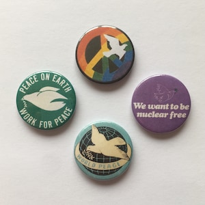 Könnte beinhalten: Vier runde Knöpfe zum Thema Frieden. Der erste Knopf ist grün mit weißem Text, der "Peace on Earth Work for Peace" lautet, und einer weißen Taube. Der zweite Knopf ist regenbogenfarben mit einem Friedenszeichen und einer weißen Taube. Der dritte Knopf ist lila mit weißem Text, der "We want to be nuclear free" lautet, und einer weißen Taube. Der vierte Knopf ist blau mit einer weißen Taube und einem Globus mit dem Text "World Peace".