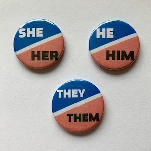 May include: Three round buttons with blue and pink halves. The top left button says "SHE HER", the top right button says "HE HIM", and the bottom button says "THEY THEM".