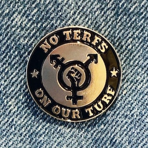 Könnte beinhalten: Eine silberne und schwarze Emaille-Anstecknadel mit dem Text "NO TERFS ON OUR FUTURE" und einem Transgender-Symbol mit einer erhobenen Faust darin.