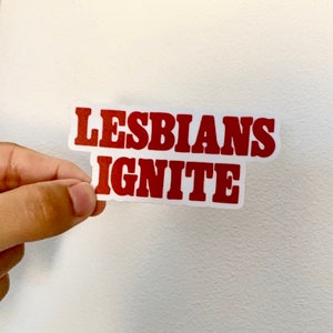 May include: A sticker with the words "LESBIANS IGNITE" in red, bold, capital letters. The text is on a white background. The sticker is being held by a person.