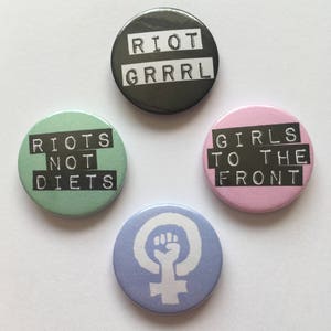 Peut inclure: Quatre boutons ronds avec des slogans et des designs différents. Le premier bouton est noir avec du texte blanc qui dit "RIOT GRRRRL". Le deuxième bouton est vert avec du texte blanc qui dit "RIOTS NOT DIETS". Le troisième bouton est rose avec du texte blanc qui dit "GIRLS TO THE FRONT". Le quatrième bouton est bleu avec un symbole féministe blanc et un poing levé.