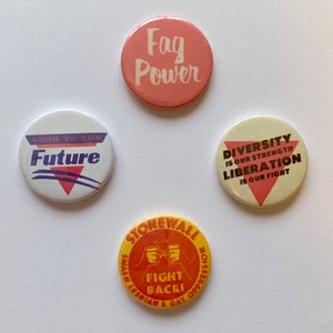 Puede incluir: Cuatro botones redondos con diferentes mensajes y diseños. El botón superior es rosa con texto blanco que dice "Fag Power". El botón de la derecha es blanco con un triángulo rosa y texto marrón que dice "Diversity is our strength Liberation is our fight". El botón de la izquierda es blanco con rayas azules y moradas y texto blanco que dice "Look to the Future". El botón inferior es amarillo con un círculo rojo y texto blanco que dice "Stonewall Smash Lesbian & Gay Oppression Fight Back!"