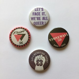 Könnte beinhalten: Vier runde Knöpfe mit verschiedenen Designs. Der erste Knopf ist rosa mit einem schwarzen Dreieck und dem Text "Pride 96". Der zweite Knopf ist lila mit dem Text "Let's Face It. We're All Queer!". Der dritte Knopf ist schwarz mit einem rosa Dreieck und dem Text "Stonewall 50 1969 2019". Der vierte Knopf ist lila mit einer Faust und dem Text "Gay Liberation Front".