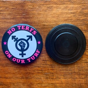Puede incluir: Un bot&oacute;n blanco y negro con texto rosa que dice "NO TERFS ON OUR TURF". El bot&oacute;n presenta un s&iacute;mbolo transg&eacute;nero con un pu&ntilde;o levantado en el centro. El bot&oacute;n est&aacute; sobre una superficie de madera.