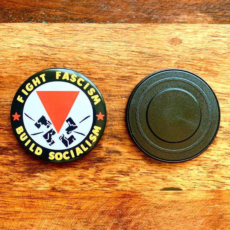 May include: A black and yellow button with the text "Fight Fascism Build Socialism" and a red triangle with a broken red arrow pointing down. The button has a black backing.