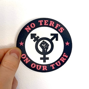May include: A round sticker with a black border and the text "NO TERFS ON OUR TURF" in pink. The center features a black transgender symbol with a raised fist. Two pink stars are on the border.