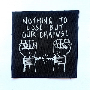 May include: Black square patch featuring white text and a graphic. The text says "NOTHING TO LOSE BUT OUR CHAINS!" with two fists breaking chains. The patch is designed to be sewn onto clothing.