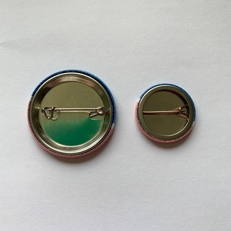 May include: Two round button pins with a blue, white, and pink striped design. The back of the pins have a metal backing with a safety pin.