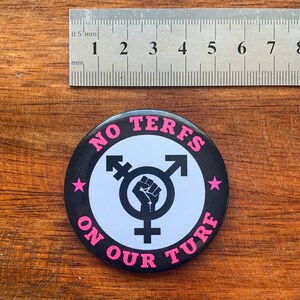 Puede incluir: Un bot&oacute;n blanco y negro con el texto "NO TERFS ON OUR TURF" en letras rosas. Un s&iacute;mbolo transg&eacute;nero con un pu&ntilde;o en alto est&aacute; en el centro del bot&oacute;n.