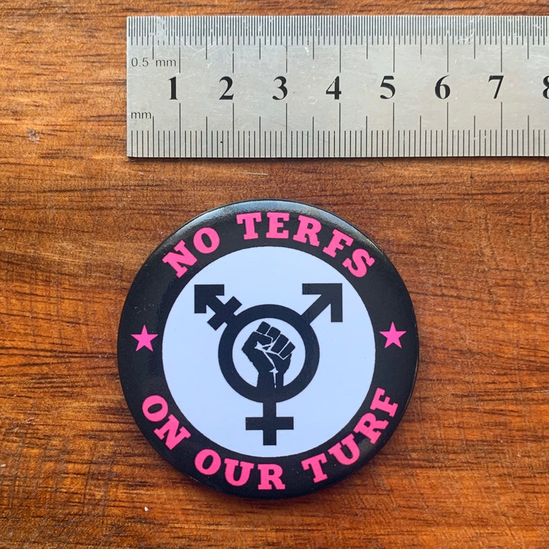 Puede incluir: Un bot&oacute;n blanco y negro con el texto "NO TERFS ON OUR TURF" en letras rosas. Un s&iacute;mbolo transg&eacute;nero con un pu&ntilde;o en alto est&aacute; en el centro del bot&oacute;n.