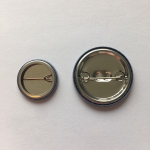 May include: Two round metal button pins with a silver finish and a blue border. The larger pin has a diameter of approximately 1.5 inches. The smaller pin has a diameter of approximately 1 inch. Both pins have a safety pin closure on the back.