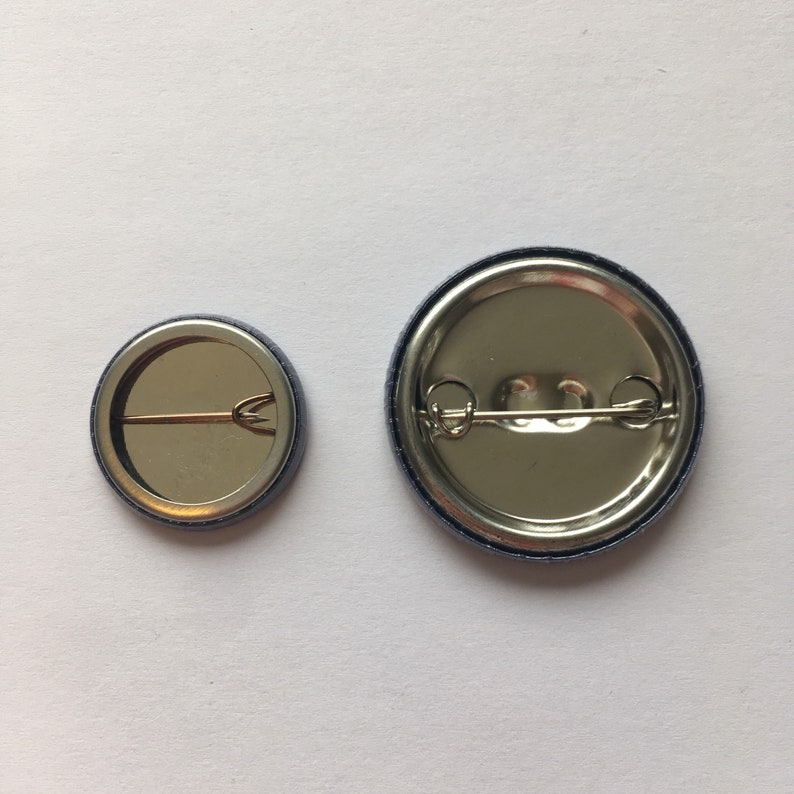May include: Two round metal button pins with a silver finish and a blue border. The larger pin has a diameter of approximately 1.5 inches. The smaller pin has a diameter of approximately 1 inch. Both pins have a safety pin closure on the back.