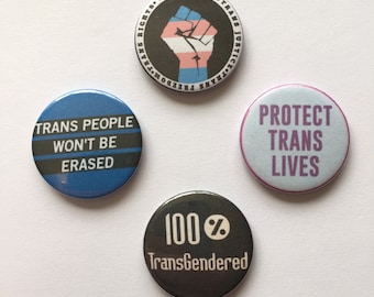 Könnte beinhalten: Vier runde Anstecknadeln mit verschiedenen Botschaften und Designs. Die erste Nadel hat einen schwarzen Hintergrund mit einer erhobenen Faust in der Mitte, mit einem Transgender-Pride-Flaggen-Design. Der Text um die Faust lautet "Trans Rights, Trans Justice, Trans Freedom, Trans Pride". Die zweite Nadel hat einen blauen Hintergrund mit weißem Text, der "Trans People Won't Be Erased" lautet. Die dritte Nadel hat einen hellblauen Hintergrund mit violettem Text, der "Protect Trans Lives" lautet. Die vierte Nadel hat einen schwarzen Hintergrund mit weißem Text, der "100% Transgendered" lautet.