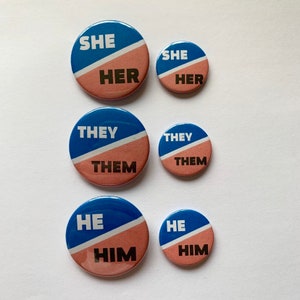 May include: Six round buttons with blue and pink halves. The buttons have the words "SHE HER", "THEY THEM", "HE HIM" in black text.
