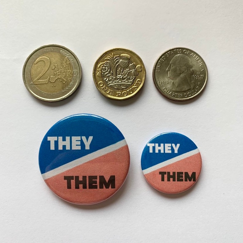 May include: Two round buttons with a blue and pink background. The text "THEY THEM" is printed in black on the buttons. The buttons are next to a US quarter, a British one pound coin, and a 2 Euro coin.
