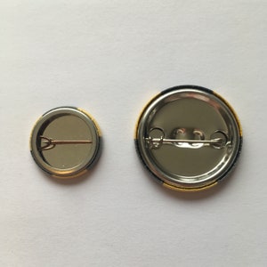 Pu&ograve; includere: Due spille rotonde in metallo con bordi neri e dorati. La spilla pi&ugrave; grande ha un diametro di circa 5 cm. La spilla pi&ugrave; piccola ha un diametro di circa 2,5 cm.