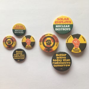 Pu&ograve; includere: Una collezione di otto bottoni rotondi con vari slogan e grafiche relative all'energia nucleare. I bottoni sono gialli, verdi, neri e rossi con testo nero. Gli slogan includono "Solar Employs Nuclear Destroys", "Women Against Nuclear Energy", "Not Nuclear Power", e "Better Active Today Than Radioactive Tomorrow".