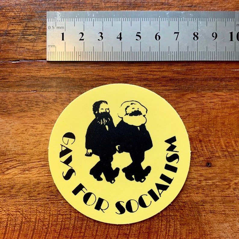 May include: A round yellow sticker with the text "GAYS FOR SOCIALISM" in black. The sticker features a black silhouette of two men walking. A ruler is placed above the sticker, measuring in millimeters.