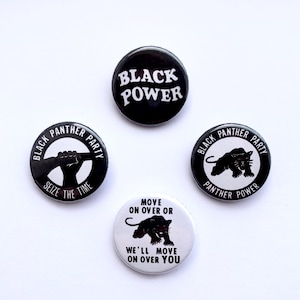 Puede incluir: Cuatro botones negros y blancos con diferentes lemas e imágenes relacionadas con el Partido Pantera Negra. Los lemas incluyen "Black Power", "Black Panther Party", "Seize the Time" y "Move on Over or We'll Move on Over You".