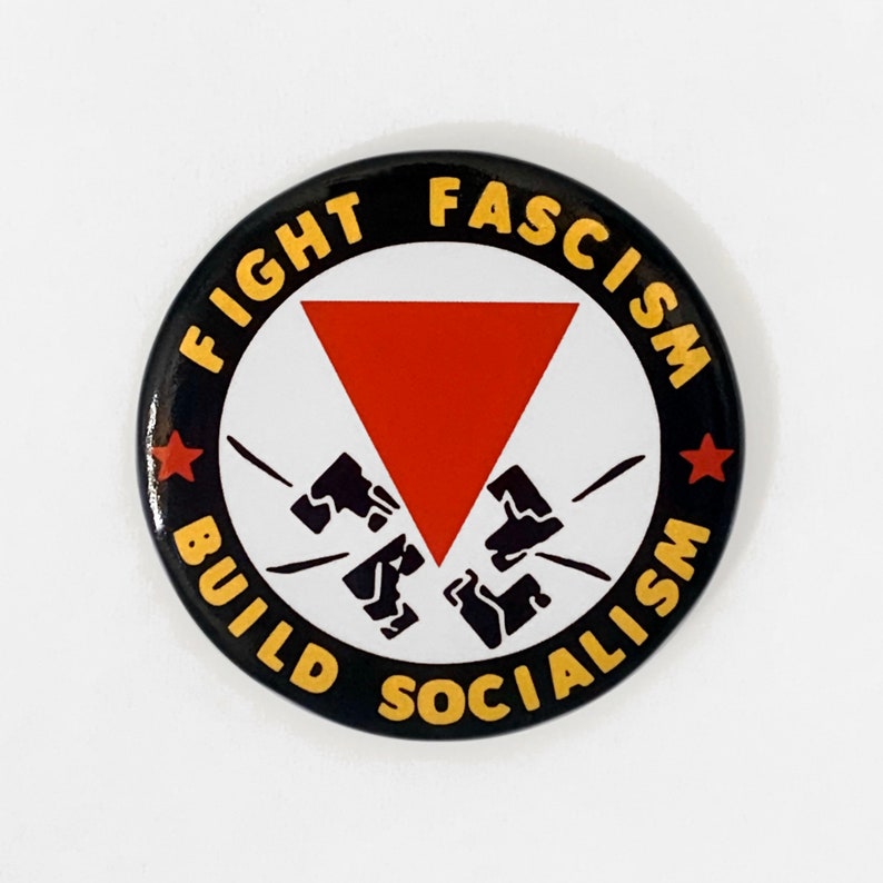 May include: Black and yellow button with the text "Fight Fascism Build Socialism" in a circle around a red triangle with black fists breaking through it.