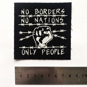 May include: A black square patch with white text and a graphic of a clenched fist. The text reads "NO BORDERS NO NATIONS ONLY PEOPLE" with barbed wire designs. A ruler is placed below the patch, measuring in millimeters.