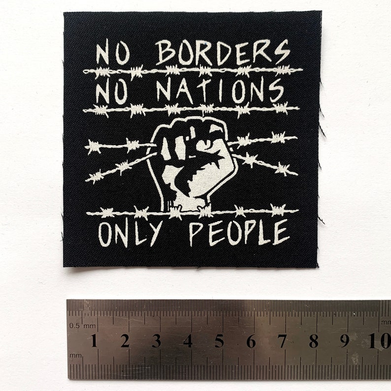 May include: A black square patch with white text and a graphic of a clenched fist. The text reads "NO BORDERS NO NATIONS ONLY PEOPLE" with barbed wire designs. A ruler is placed below the patch, measuring in millimeters.