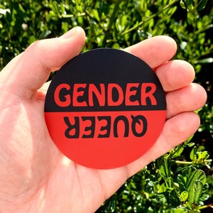 May include: A black and red round button with the text "GENDER QUEER" in white.