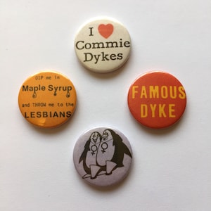 Może przedstawiać: Cztery odznaki z różnymi wzorami. Jedna odznaka jest biała z czerwonym sercem i tekstem "I Commie Dykes". Inna odznaka jest pomarańczowa z tekstem "DIP me in Maple Syrup and THROW me to the LESBIANS". Trzecia odznaka jest czerwona z tekstem "FAMOUS DYKE". Ostatnia odznaka jest fioletowa z dwoma pingwinami trzymającymi się za ręce i symbolem żeńskim.