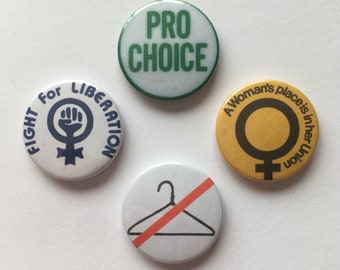 May include: Four round buttons with different messages and symbols. The first button is white with blue text that says "Fight for Liberation" and a blue female symbol with a raised fist. The second button is white with green text that says "Pro Choice". The third button is yellow with black text that says "A Woman's place is in her Union" and a black female symbol. The fourth button is white with a gray hanger and a red line through it.