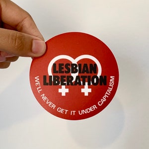May include: A red circle sticker with the text "LESBIAN LIBERATION" in black and white female symbols. The text "WE'LL NEVER GET IT UNDER CAPITALISM" is written in a circle around the outside.